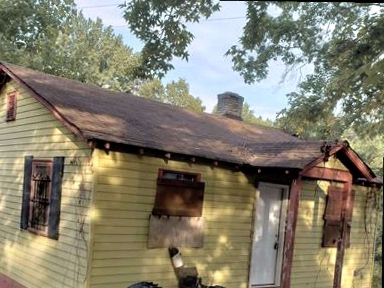 Cottage before renovation — failing roof, damaged siding, boarded window, debris in the yard